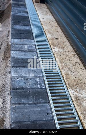 Vue rapprochée d'une grille de drainage métallique nouvellement installée à côté de pavés sombres et d'une surface en béton à l'extérieur d'une porte de garage. Banque D'Images