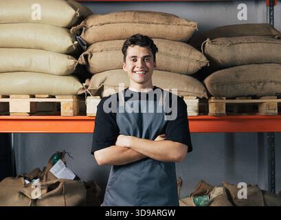 Portrait d'un jeune ouvrier masculin confiant dans un tablier à l'entrepôt. Homme aux bras croisés portant un tablier debout à l'entrepôt. Portrait de jeune confiant Banque D'Images