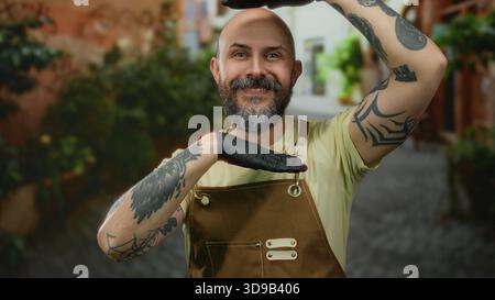 Homme chauve barbu en salopette avec tatouages et gants pose de manière créative à l'extérieur sur une rue pittoresque de la ville. Banque D'Images