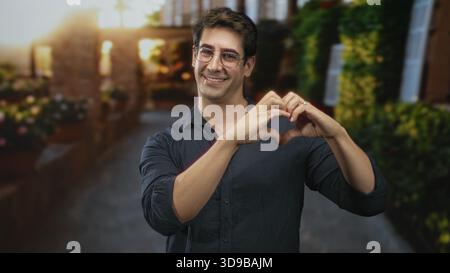 Homme avec des lunettes forme la forme de coeur avec les mains tout en souriant sur une rue ensoleillée bordée de jardinières et de verdure ; affection de connexion. Banque D'Images