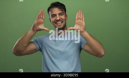 Jeune homme hispanique souriant sur fond vert isolé dans une chemise décontractée faisant des gestes d'encadrement avec les mains, mettant en valeur l'expression joyeuse et po Banque D'Images