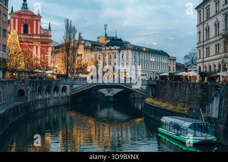 Rivière festive de la ville avec des bâtiments illuminés, des arbres décorés et des reflets brillants au crépuscule. Ljubljana, Slovénie Banque D'Images