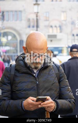 Un homme d'âge moyen se tient debout dans une rue italienne concentrée sur son smartphone portant des lunettes et une veste matelassée sombre. Banque D'Images
