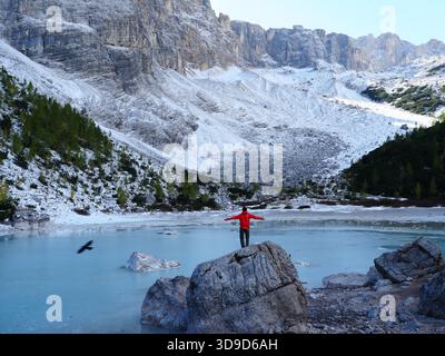 Une personne debout sur un rocher avec les bras tendus face au Lago di Sorapis partiellement gelé et les sommets Dolomites environnants au début de l'hiver, Italie. Banque D'Images