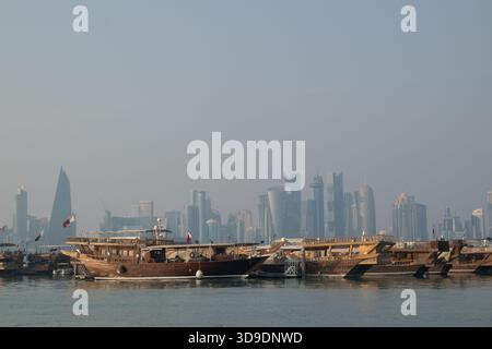Des boutres en bois sont ancrés dans une marina avec l'horizon de Doha s'élevant en arrière-plan tandis que la lumière du matin projette une douce lueur. Banque D'Images