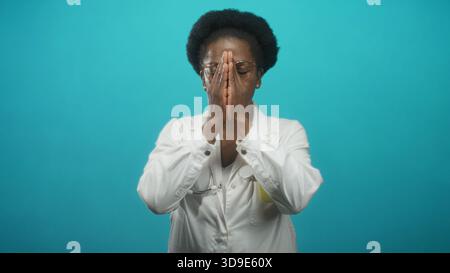 Femme médecin en blouse de laboratoire blanche et stéthoscope, portant des lunettes, des tasses mains sur le visage puis ouvre les paumes sur fond de studio turquoise ; calme resur Banque D'Images