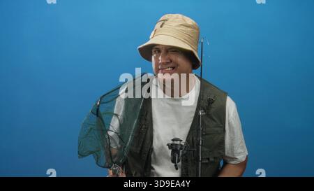 Homme pêcheur portant chapeau seau et gilet de pêche tient un filet d'atterrissage et une canne, souriant dans le studio ; contentement de loisirs. Banque D'Images