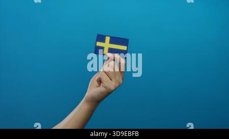 Homme tenant le drapeau suédois brodé sur fond bleu isolé, mettant en valeur l'identité nationale avec l'accent sur les détails de la main et les riches symboli culturels Banque D'Images