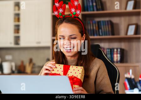 Joyeuse femme tenant une boîte-cadeau tout en ayant une réunion virtuelle de Noël. Banque D'Images