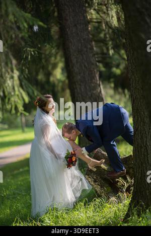 Groom embrasse doucement le pied de la mariée comme elle se tient sur l'herbe à côté de grands pins, mariée tenant bouquet - une scène de mariage ludique mais tendre baignée dans s. Banque D'Images