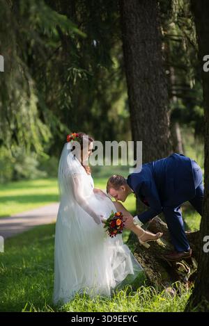 Groom embrasse doucement le pied de la mariée comme elle se tient sur l'herbe près des pins, mariée tenant un bouquet vibrant dans la robe de mariée - un mariage ludique mais tendre Banque D'Images