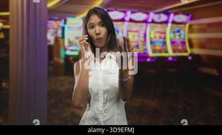 Jeune femme thaïlandaise tenant smartphone lève la main ouverte pour s'arrêter près des machines à sous colorées dans le bâtiment ; salutation joyeuse. Banque D'Images