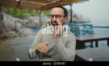 Homme mûr criant au bord de la mer avec café en plein air en arrière-plan, mettant en valeur une atmosphère de plage animée et une communication expressive. Banque D'Images