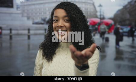 Femme souriant avec les cheveux bouclés étend sa main vers l'avant sur une rue humide flanquée de bâtiments historiques ; invitation. Banque D'Images