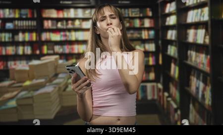 Une jeune femme en débardeur rose se mord l'ongle tout en tenant un smartphone au milieu de livres empilés et d'étagères dans une bibliothèque ; anxiété. Banque D'Images