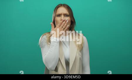 Jeune femme blonde couvre la bouche dans la surprise sur fond vert, exprimant l'étonnement avec sa main délicatement placée sur ses lèvres, transportant Banque D'Images