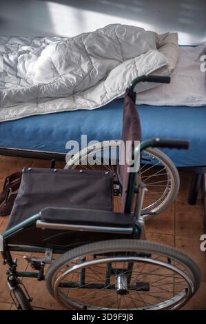 Fauteuil roulant positionné à côté d'un lit non fait dans un salon calme de soins de santé ou assisté. L'image véhicule les thèmes du handicap, de l'aide à la mobilité, Banque D'Images