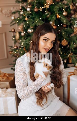 Une femme aux cheveux longs sourit tout en tenant doucement un petit lapin. Elle est entourée de cadeaux et d'un magnifique sapin de Noël orné d'ornements Banque D'Images