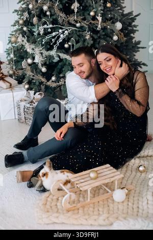 Un couple est assis sur un tapis moelleux près d'un sapin de Noël décoré, souriant et partageant un moment joyeux. Devant eux, un lapin explore un petit sle en bois Banque D'Images