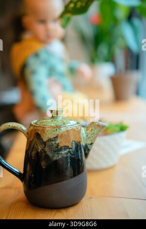 Table avec théière et nourriture servie dans un café lumineux. Enfant en bas âge assis en arrière-plan créant une atmosphère familiale. Parentalité, amour et bonheur dans un restaurant public. || autorisation du modèle Banque D'Images