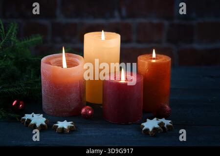 Quatre bougies orange et rouge apportant une lumière chaude dans la nuit sombre pour Noël, l'Avent ou le nouvel an, quelques biscuits, boules et branches de sapin contre un mur de briques, foyer sélectionné, profondeur de champ étroite Banque D'Images