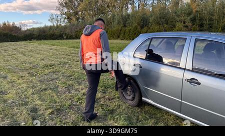 Un mécanicien masculin caucasien dans une veste orange testant la roue arrière droite d'une voiture argentée en la poussant avec son pied tout en se tenant sur une route herbeuse Banque D'Images