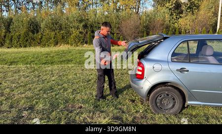 Un mécanicien masculin caucasien dans une veste orange et grise ouvrant le coffre d'une voiture argentée de la vue de côté tout en se tenant debout sur l'herbe verte sous l'arrière Banque D'Images