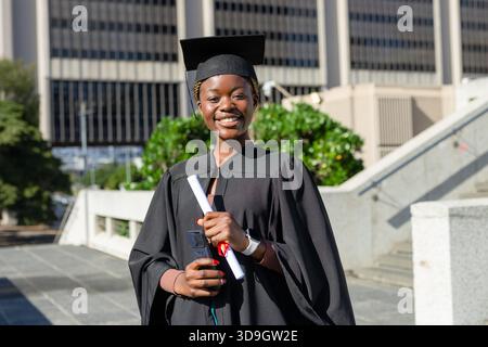 Femme afro-américaine en casquette et robe debout sur la place de l'université détenant le diplôme, smartphone Banque D'Images