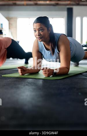 Diverses amies féminines tenant la planche d'avant-bras dans le studio de fitness sur des tapis verts portant smartwatch Banque D'Images
