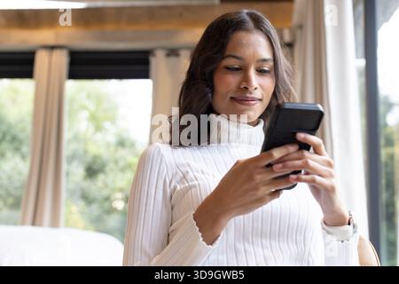 Femme adulte asiatique assise à côté d'une grande fenêtre dans le salon tenant smartphone et portant un pull Banque D'Images