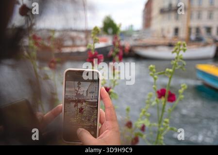 Les mains d'une femme sont visibles pendant qu'elle prend une photo des fleurs devant elle avec de l'eau, des bateaux et des bâtiments en arrière-plan Banque D'Images