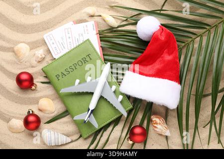 Vacances de Noël. Passeport avec billets, modèle d'avion, chapeau de Père Noël, coquillages, feuille de palmier et ornements sur la plage, pose à plat Banque D'Images