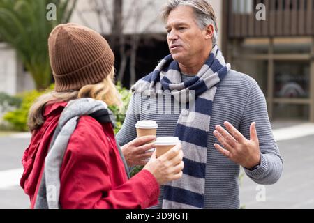 Amis d'âge moyen portant des foulards et un bonnet debout dans la cour tenant des tasses à café, faisant des gestes Banque D'Images