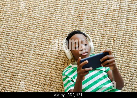 Enfant afro-américain couché sur un tapis tissé à la maison tenant un smartphone portant des écouteurs, espace de copie Banque D'Images