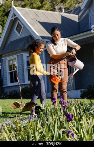 Mère asiatique et fille afro-américaine arrosant lit d'iris violet avec arrosoirs dans la cour avant Banque D'Images