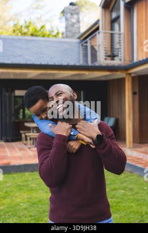 Père afro-américain portant son fils sur le dos riant dans la cour avant avec table de patio et smartwatch Banque D'Images