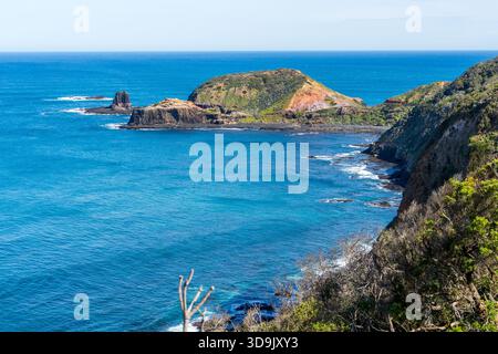 Les eaux bleues vibrantes de l'océan rencontrent un littoral accidenté orné de collines verdoyantes et de formations rocheuses sombres sous un ciel clair et lumineux. Banque D'Images