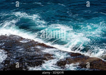 Les vagues turquoises éclatantes de l'océan s'écrasent contre un littoral rocheux sombre et accidenté, créant de la mousse blanche et un mouvement dynamique de l'eau. Banque D'Images