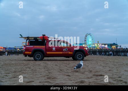 Santa Monica, CALIFORNIE, États-Unis. 26 septembre 2025. - Un camion Toyota Tacoma rouge de sauveteur du comté de Los Angeles avec planches de surf est assis sur le sable, une mouette à proximité Banque D'Images