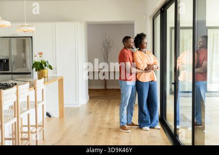 Couple afro-américain debout par des portes vitrées coulissantes dans une cuisine moderne avec îlot, espace copie Banque D'Images