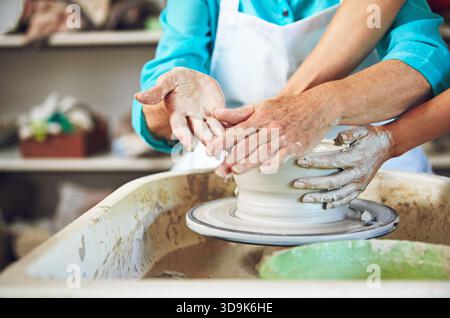 Poterie, classe et mains dans l'atelier avec l'aide, roue d'argile et aide de l'enseignant dans le passe-temps créatif. Art, personnes et instructeur en studio avec soutien Banque D'Images