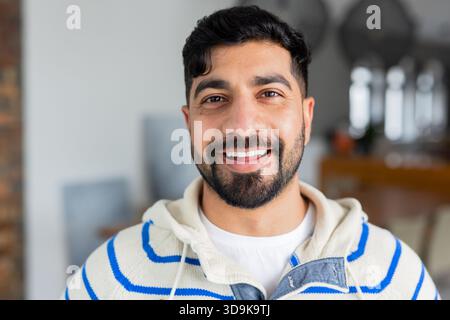 Homme indien debout souriant dans la cuisine de la maison portant un sweat à capuche rayé par comptoir avec des casseroles suspendues Banque D'Images