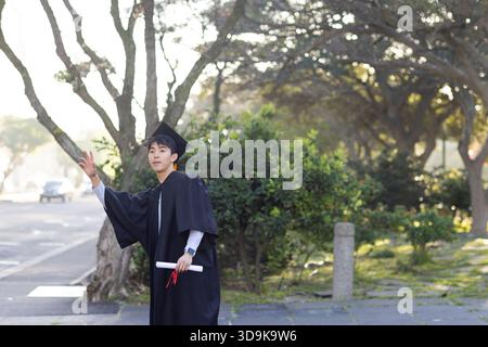 Homme asiatique debout sur la passerelle à côté de la route portant la robe de graduation et mortarboard tenant le diplôme Banque D'Images
