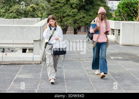 Diverses amies féminines marchant sur la place du campus discutant au téléphone, tenant des cahiers et des sacs Banque D'Images