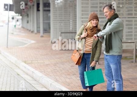Couple senior vérifiant le temps sur la montre-bracelet tenant le sac à provisions et le sac à main à l'espace de copie du poste Banque D'Images