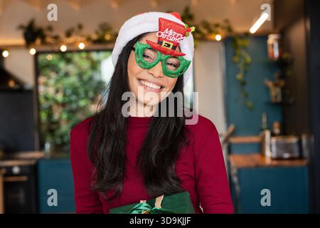 Femme mi-adulte portant chapeau de Père Noël et lunettes debout dans la cuisine avec des lumières, tenant une boîte cadeau Banque D'Images
