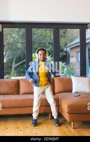 Enfant mâle noir dansant dans le salon à côté du canapé près des portes vitrées, portant des écouteurs Banque D'Images
