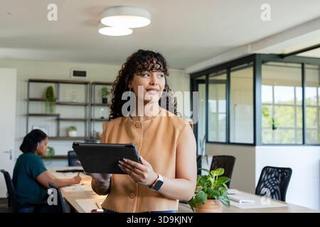 Collègues afro-américaines debout tenant une tablette portant un chemisier bronzé et travaillant au bureau Banque D'Images