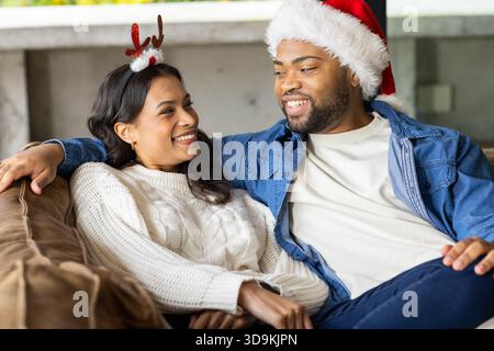 Couple diversifié assis sur le canapé près de la cheminée à la maison portant chapeau de Père Noël, serre-tête en bois de renne Banque D'Images