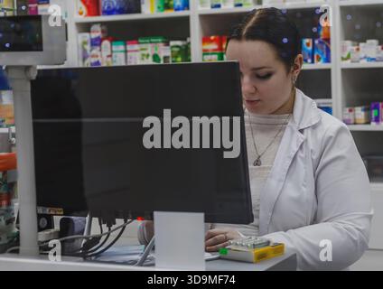 La photo montre une pharmacienne en blouse blanche traitant un achat de médicaments à un ordinateur, entourée d'étagères remplies de produits pharmaceutiques p Banque D'Images